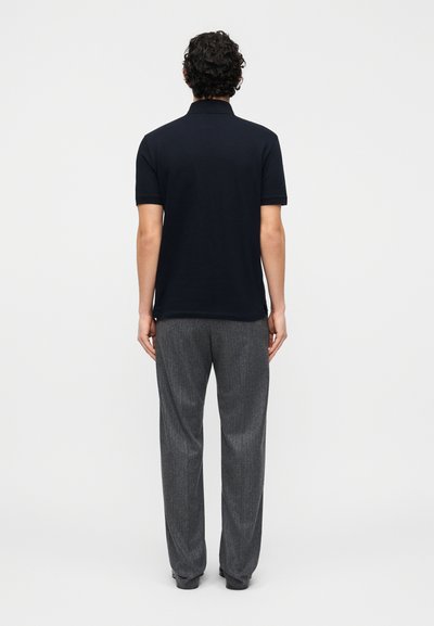 Άνδρας με κατσαρά μαλλιά στέκεται πλάτη, φορώντας μαύρο κοντομάνικο πόλο μπλουζάκι και γκρι παντελόνι φαρδύ με ίσια γραμμή, μπροστά σε απλό φόντο.