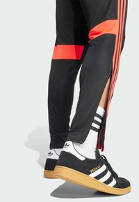 Pantalones atléticos negros con franjas rojas, con cremalleras en los laterales; combinados con zapatillas Adidas en blanco y negro con suela de goma.