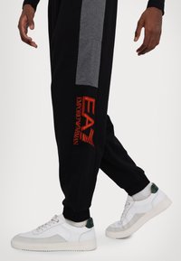 Pantaloni jogger neri con pannello laterale grigio, caratterizzati dal logo rosso "EA7 Emporio Armani". Abbinati a sneakers bianche con un accento verde sul tallone.