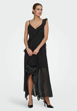 Femeie purtând o rochie neagră asimetrică cu volane și bretele subțiri, ținând o mică geantă neagră și purtând pantofi cu toc negri.