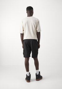 Light beige cotton t-shirt and black cargo shorts, featuring side pockets. Worn with black boots and white socks, seen from the back.