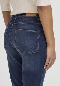 Vaqueros de mezclilla azul oscuro con una cintura de altura media, que cuentan con dos bolsillos traseros y una marca de cuero marrón en la cinturilla. Textura suave.