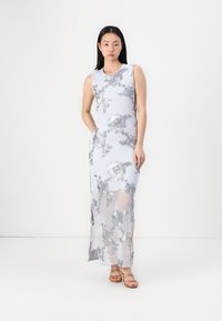 Robe longue ajustée en tissu blanc transparent avec des motifs floraux noirs. Présente un design sans manches et des fentes sur les côtés ; associée à des sandales beiges.