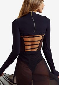 Kobieta nosząca dopasowaną czarną bluzkę z długimi rękawami i otwartym tyłem, z poziomymi paskami, połączoną z przezroczystą czarną spódnicą na czarnych majtkach.