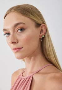Goudkleurige oorbellen met een groene edelsteen en een decoratief gebogen ontwerp. Het model draagt een roze haltertop en heeft lang, steil haar.