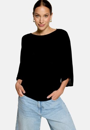 Femme portant un haut noir à manches longues larges, un jean bleu clair, les mains dans les poches et des boucles d'oreilles créoles dorées.