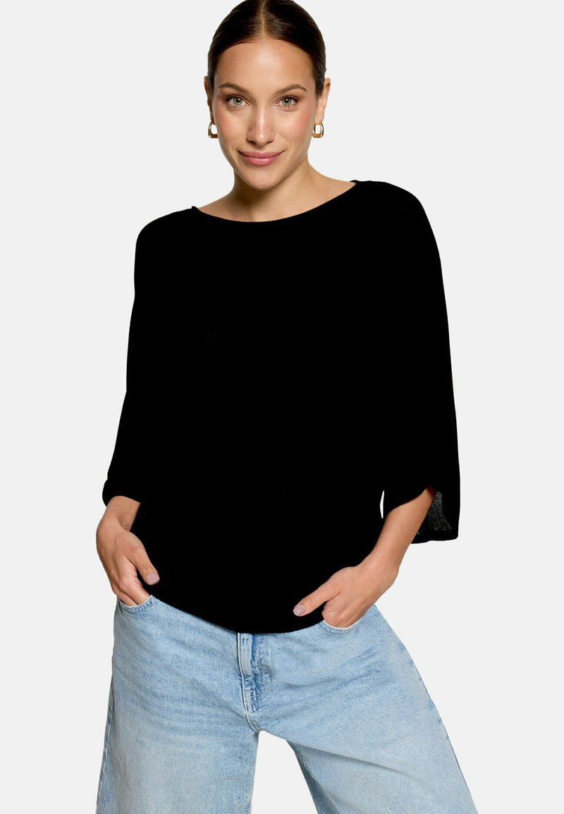Femme portant un haut noir à manches longues larges, un jean bleu clair, les mains dans les poches et des boucles d'oreilles créoles dorées.
