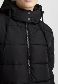 Veste noire rembourrée avec un col montant, fermeture éclair et cordon ajustable. Présente un design matelassé et des boutons-pression.