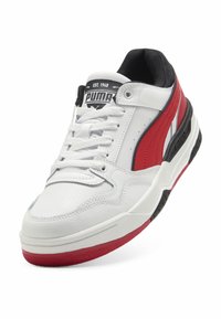 Białe sneakersy z czerwonymi akcentami i czarnymi detalami. Posiadają teksturowaną powierzchnię, wyściełany kołnierz oraz gumową podeszwę z wyraźnymi wzorami zapewniającymi przyczepność.