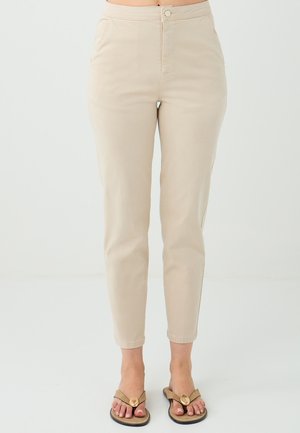 Beige slim-fit broek gedragen met slippers, welke een casual outfit toont tegen een effen witte achtergrond. Focus op de onderkant van het lichaam.
