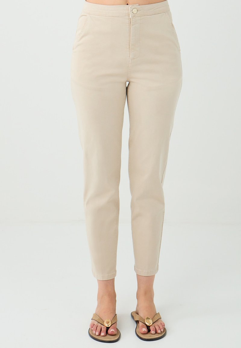 Beige slim-fit broek gedragen met slippers, welke een casual outfit toont tegen een effen witte achtergrond. Focus op de onderkant van het lichaam.