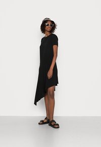 Vestido preto assimétrico com mangas curtas, tecido suave e uma bainha que se inclina de forma acentuada. Combinado com sandálias pretas e óculos de sol.