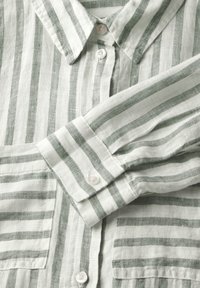 Camicia di lino a righe verde chiaro con bottone frontale, colletto e maniche lunghe con polsini a bottone. Tessuto con texture visibile.