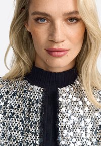 Black knitted turtleneck under a textured, multicolored jacket featuring black, silver, and white sequins, with a fitted silhouette.