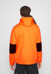 Felpa con cappuccio in morbido pile arancione brillante, con accenti in velluto nero sulle maniche. Presenta un tascone a marsupio e piccole grafiche di uccelli sulla spalla.