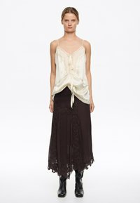 Kobieta w kremowym jedwabnym topie z ramiączkami i wiązaniem, ciemnobrązowej spódnicy midi z koronkową lamówką oraz czarnych skórzanych botkach na jednolitym białym tle.