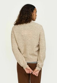 Beżowy sweter dziany o teksturowanym wykończeniu, luźny krój i ściągacze; noszony z brązowymi spodniami, widok z tyłu.