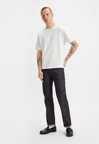 Camiseta blanca de algodón, corte relajado, mangas cortas. Jeans oscuros, de pierna recta, con costuras sutiles. Mocasines negros con suela visible.