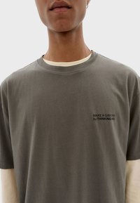 Graues Baumwoll-T-Shirt mit Rundhalsausschnitt, das eine weiße Innenschicht und schwarzen Text auf der Vorderseite trägt: "EINEN GARTEN MACHEN mit THINKING MU."