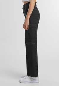 Pantaloni cargo neri con vestibilità dritta, dotati di molteplici tasche e cerniere, realizzati in tessuto resistente, abbinati a sneakers bianche.