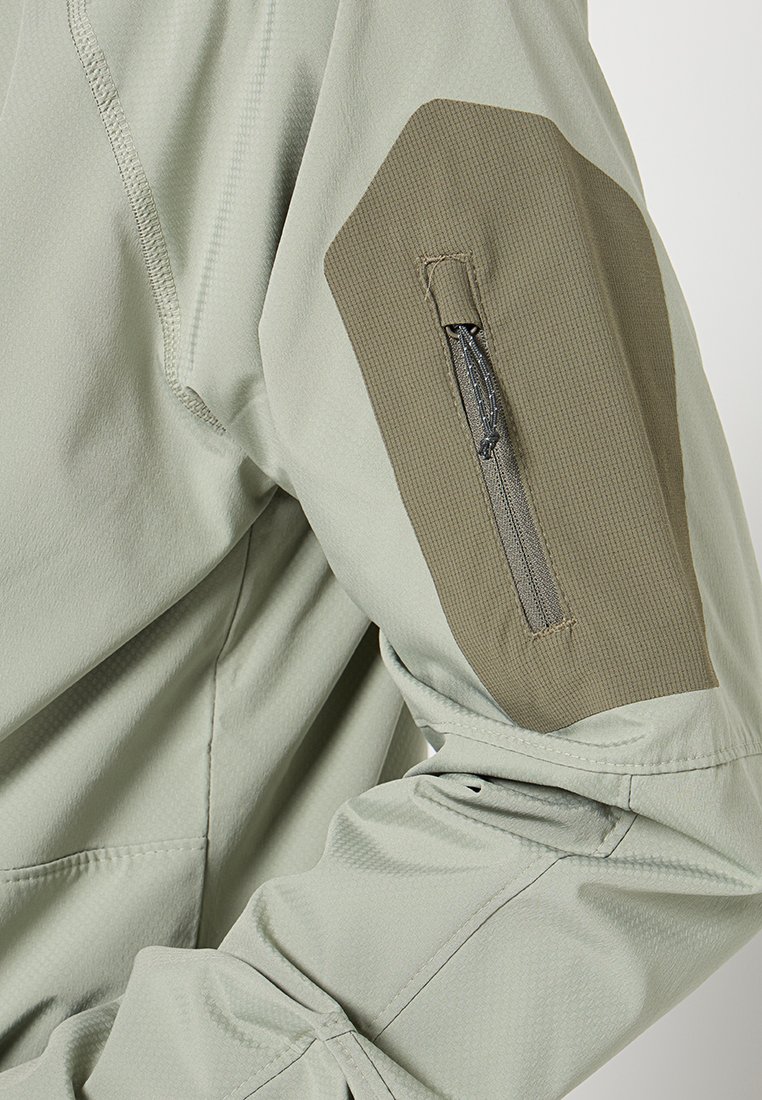 Hellgrüne Jacke mit einem strukturierten, dunkelgrünen Ellbogenpatch. Verfügt über eine Reißverschlusstasche am Ärmel und glattes Material insgesamt.