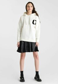 Witte hoodie met zwart logo, gecombineerd met een zwarte neplederen flare rok. Model draagt zwarte textuursokken en glanzende zwarte schoenen.