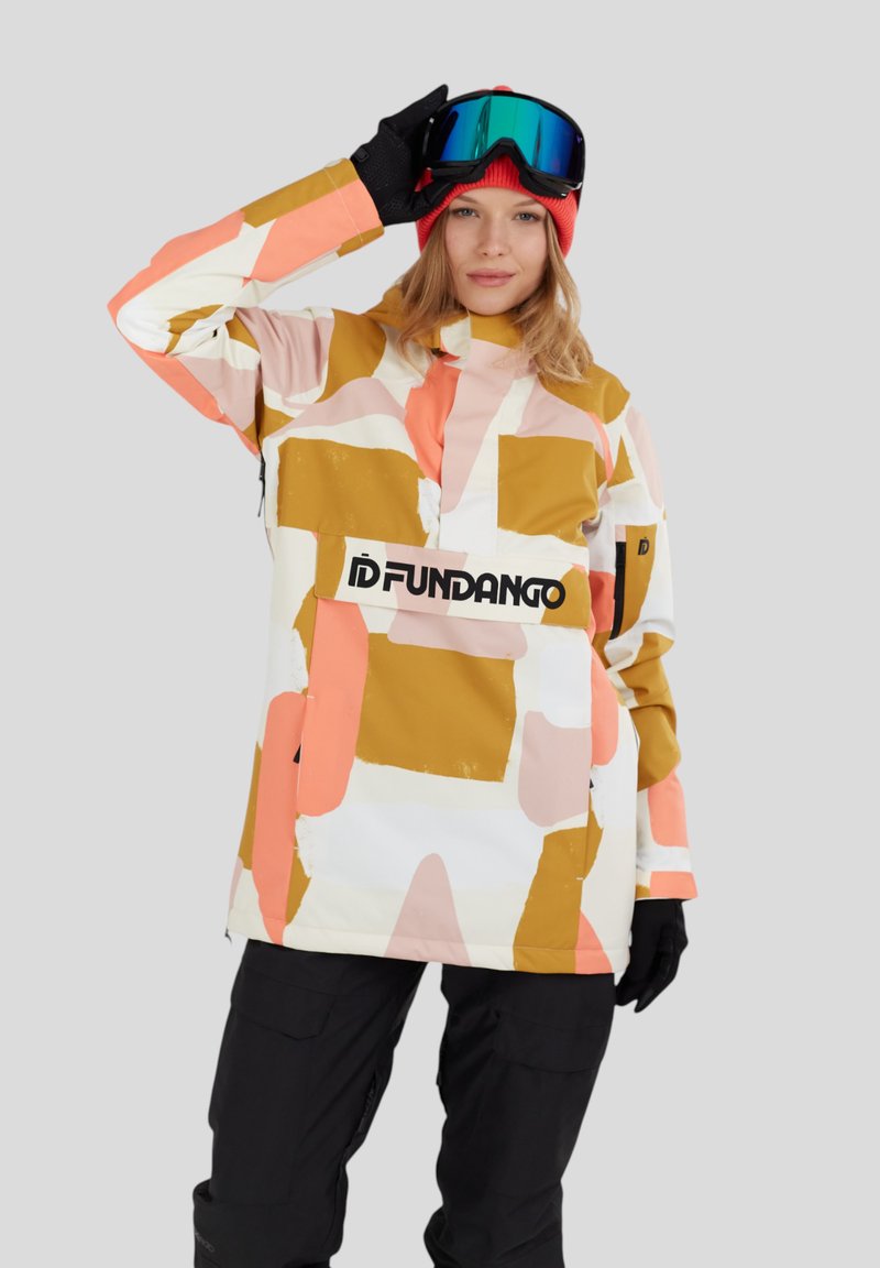 Femme portant une veste d'hiver multicolore à motifs, un bonnet rouge, des gants noirs, un pantalon noir et des lunettes de ski bleues, ajustant ses lunettes avec la main.