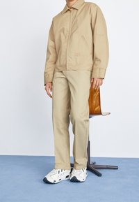 Beige bunda na zip s límcem, spárovaná s odpovídajícími béžovými kalhotami a bílými teniskami. Hladká textura, minimalistický design a jemné značení.