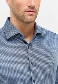 Uomo con barba corta che indossa una camicia blu a fantasia con colletto aperto, su sfondo semplice.