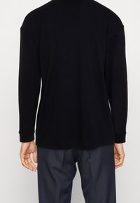 Henkilö, jolla on päällään yksinkertainen musta pitkähihaisena paita ja tummat raitalahkeet, näkyy selkä menosuunnassa neutraalin taustan edessä.