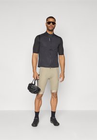 Gilet de cyclisme sans manches noir au design ajusté, associé à un short beige. Les accessoires comprennent un casque noir et des lunettes de soleil. Chaussures de cyclisme noires.