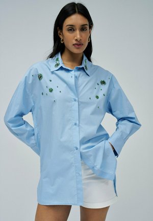 Chemise boutonée bleu clair avec broderie florale verte sur le col et le corps ; possède des manches longues et un design décontracté.
