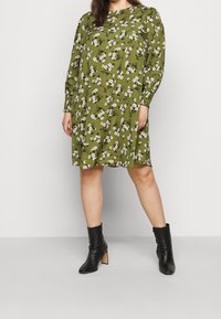 Abito floreale verde con maniche lunghe, svasato e con scollo rotondo. Abbinato a stivaletti neri alla caviglia con tacco a blocco.