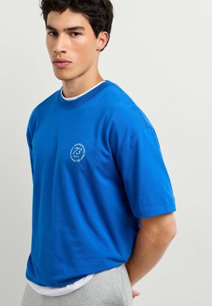 Junger Mann mit kurzen dunklen Haaren, der ein hellblaues T-Shirt mit kleinem weißem rundem Logo auf der Brust trägt und vor einem schlichten hellen Hintergrund steht.