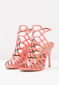 Öppna tåhögklackade skor med geometrisk utskärning, gjorda av glittrigt korallmaterial, med stiletthäl och vristrem.