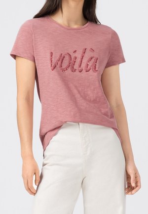 Mujer con camiseta rosa de manga corta con texto de lentejuelas "voilà" y pantalones blancos, mostrada desde el cuello hasta las caderas sobre un fondo liso.