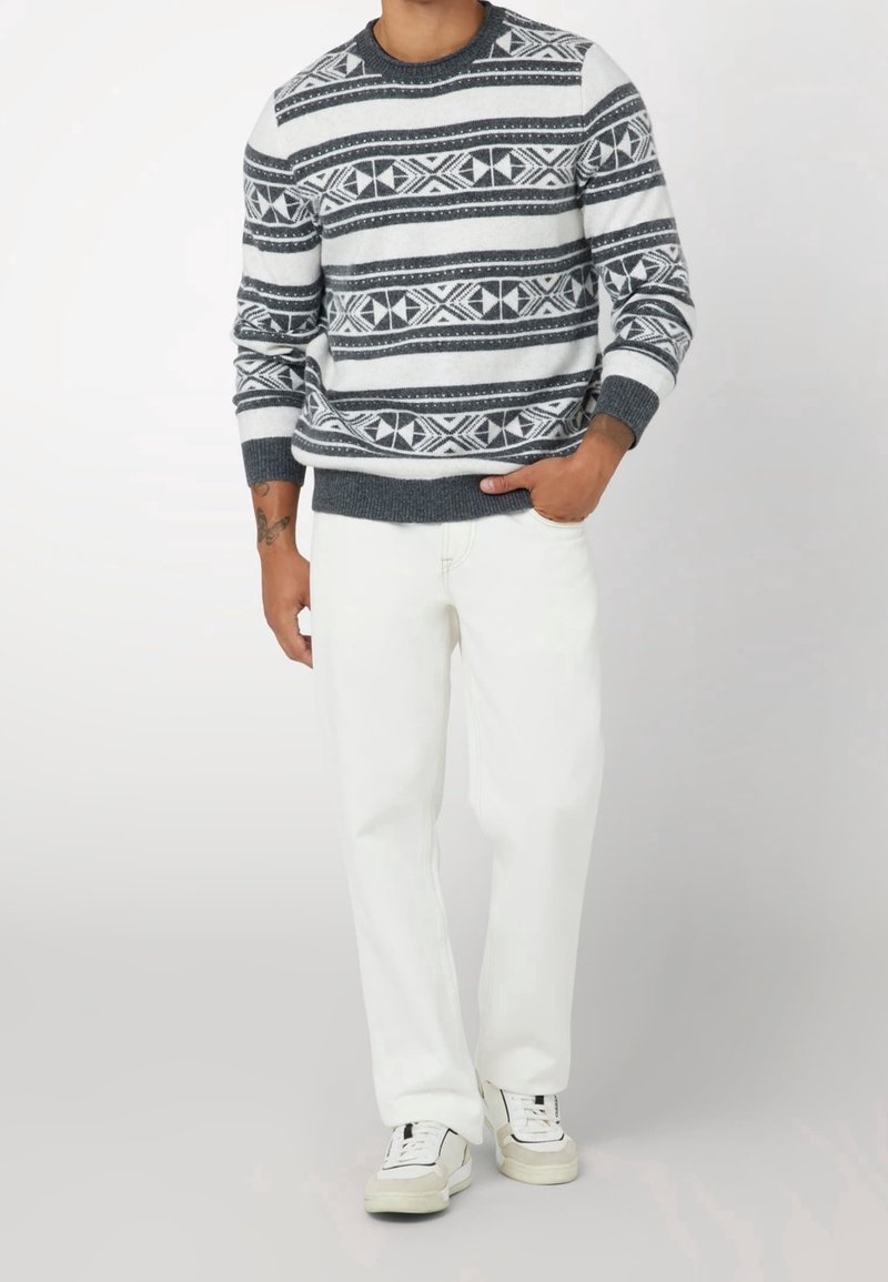 Homme portant un pull gris et blanc à motifs, un pantalon blanc et des baskets blanches, debout avec une main dans la poche.