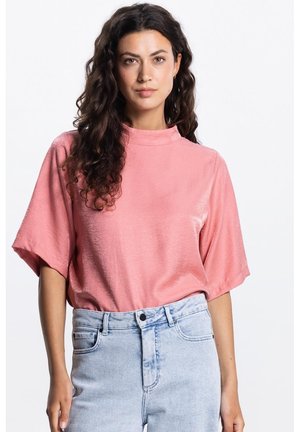 Vrouw met lang krullend haar, gekleed in een loszittend roze topje met korte mouwen, ingestopt in lichtblauwe, hooggetailleerde spijkerbroek, staand tegen een witte achtergrond.