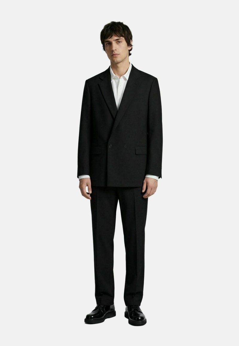 Joven de pie vestido con un traje negro cruzado, camisa blanca y zapatos de vestir negros sobre un fondo liso y claro.