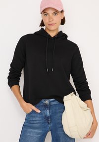 Sudadera negra con capucha y cordones, combinada con unos pantalones vaqueros azules. El modelo sostiene un bolso color crema. Una gorra de pana rosa completa el look.