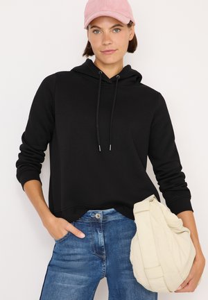 Sudadera negra con capucha y cordones, combinada con unos pantalones vaqueros azules. El modelo sostiene un bolso color crema. Una gorra de pana rosa completa el look.