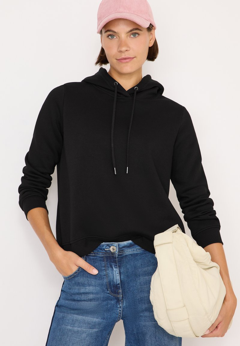 Sudadera negra con capucha y cordones, combinada con unos pantalones vaqueros azules. El modelo sostiene un bolso color crema. Una gorra de pana rosa completa el look.