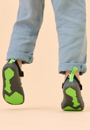 Kind trägt hochgekrempelte hellblaue Hose und schwarze Schuhe mit leuchtend grünen Sohlen, geht auf einer hellbeigen Oberfläche.
