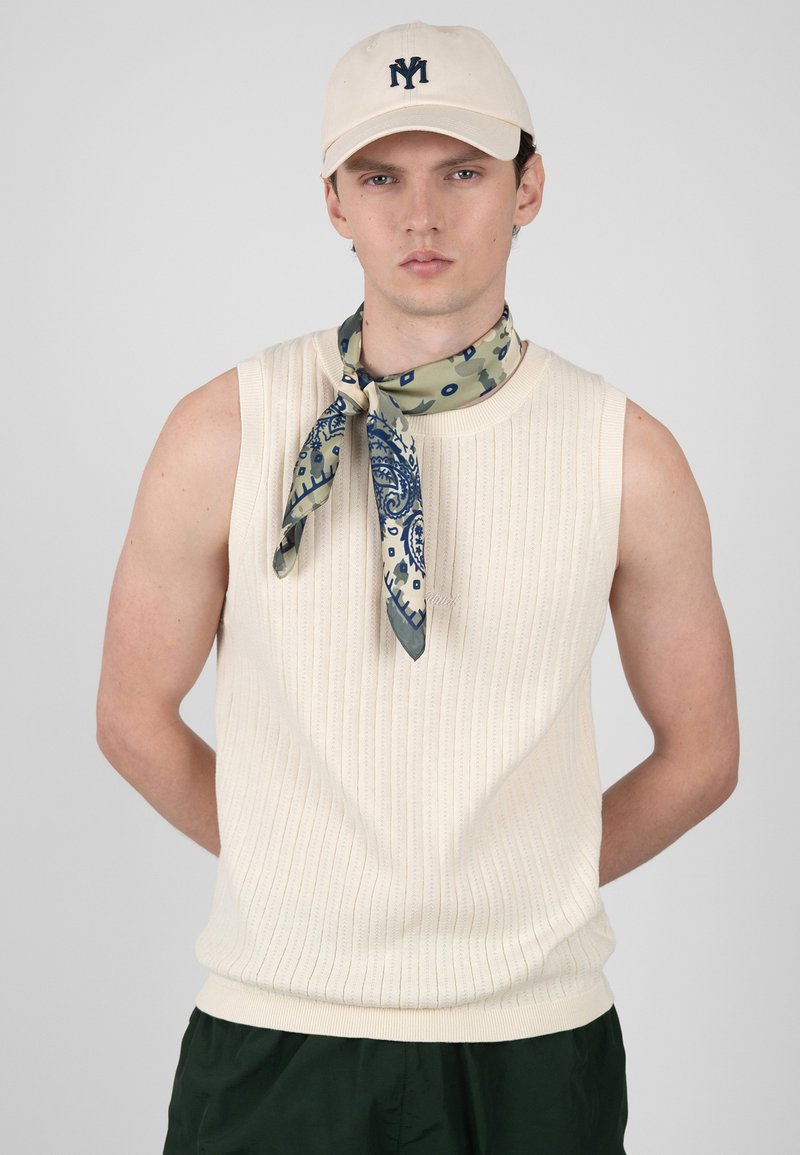 Jeune homme portant un haut en maille sans manches crème, un foulard à motifs autour du cou, une casquette beige avec un logo foncé et un pantalon vert foncé, sur un fond uni.