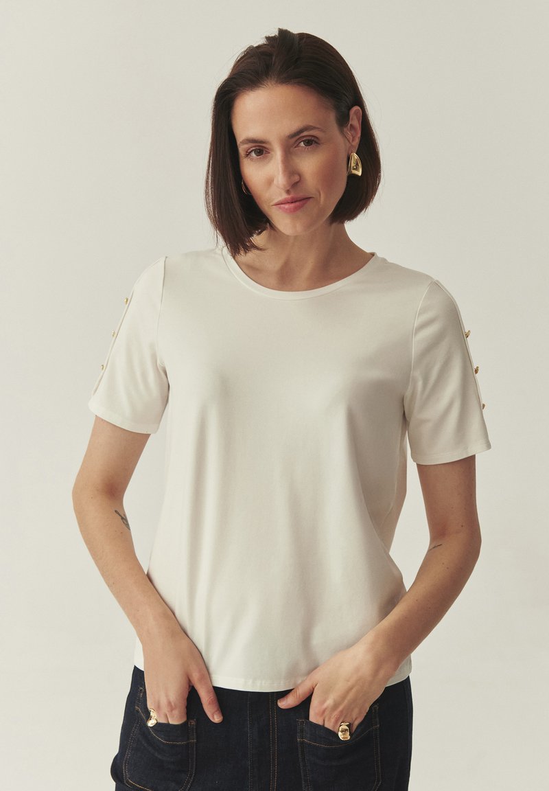 T-shirt blanc à manches courtes avec un col rond, agrémenté de boutons dorés sur les épaules. Tissu lisse et coupe décontractée.