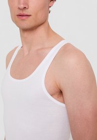 Vit bomullstopp med breda axelband, rund halsringning och figurnära design, bärs av en person från axlarna och upp.