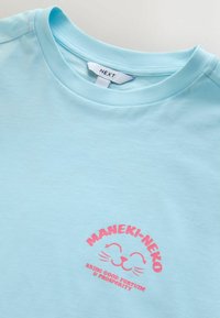 Svetlomodra bombažna majica z roza potiskom mačjega obraza in napisom "MANEKI-NEKO PRINAŠA DOBRO SREČO IN BLAGOSTANJE." Okrogel izrez.