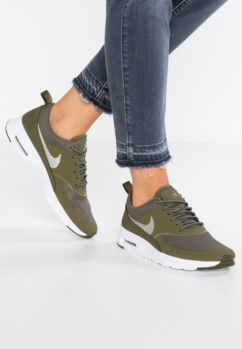Pieds chaussés de baskets Nike vert olive et blanches associées à un jean court gris foncé effiloché sur fond blanc.