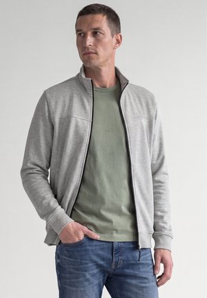 Homme portant une veste zippée gris clair sur un t-shirt vert avec un jean bleu, debout avec une main dans la poche devant un fond uni.