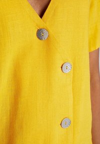 Žlutý lněný top s designem do V, krátkými rukávy a třemi kulatými stříbrnými knoflíky s texturovaným povrchem.